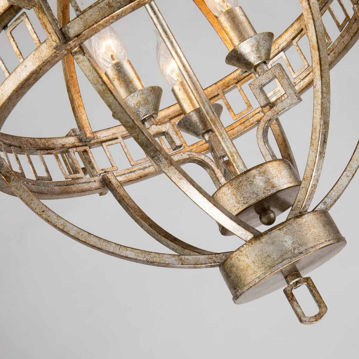 Close-up of the Lemuria 3 Light Globe Chandelier, showcasing its ornate metallic design with a spherical frame. The chandelier features intricate geometric detailing and exposed bulbs, while its gold finish highlights a Grecian key motif, beautifully enhancing the elegance of its ivory shade against a neutral background.
