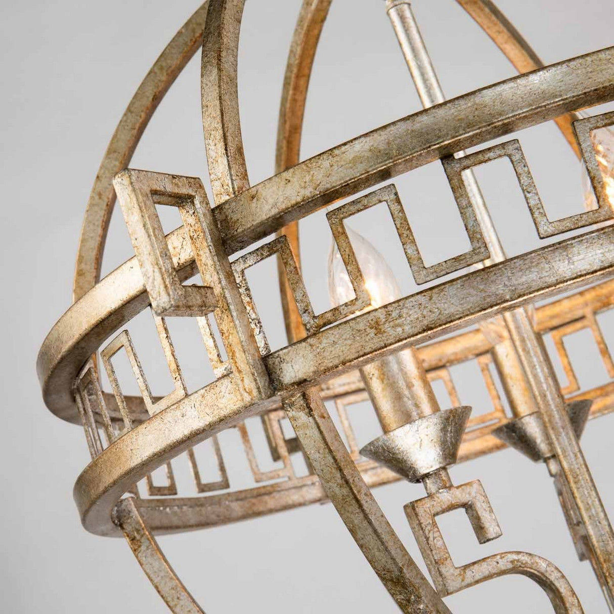 A close-up view of the Lemuria 3 Light Globe Chandelier reveals its spherical design featuring a Grecian key motif. The gold finish imparts a vintage, rustic charm, with small candle-like bulbs nestled within its structure. Set against a plain, light gray background, it highlights the fixture's timeless elegance and style.