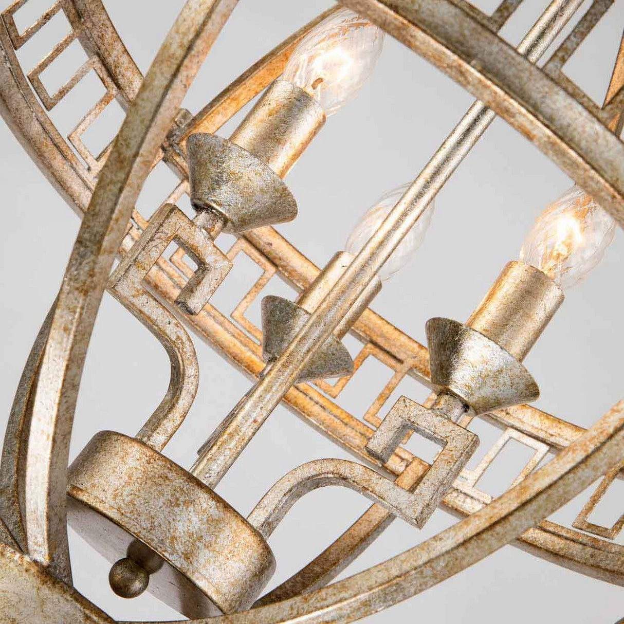 A close-up of the Lemuria 3 Light Globe Chandelier - Gold With Ivory Shade highlights its ornate metallic design featuring intricate geometric patterns, including a Grecian key motif. The chandelier's distressed gold finish adds timeless elegance, and the adjustable rods allow for customizable height to suit any space.