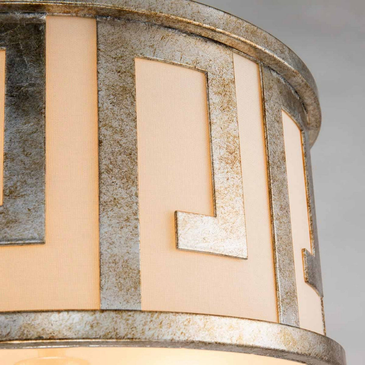 A close-up of the Lemuria 2 Light Duo-Mount Medium Pendant/Semi-Flush reveals its decorative cylindrical shade featuring a silver and beige geometric pattern reminiscent of a Grecian key motif. Its soft lighting casts a warm ambiance, accentuating the elegant metallic finish and angular design in gold with an ivory shade.