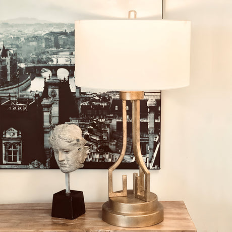 A Lemuria 1 Light Table Lamp with a gold finish and ivory shade sits elegantly on a wooden surface. Next to it is a sculpture of a head on a black pedestal, adorned with intricate Grecian key motif details. In the background, a black-and-white cityscape photograph completes the aesthetic tableau.