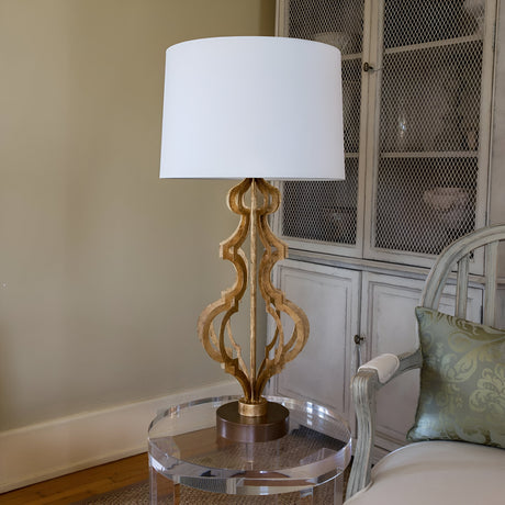 An Octavia 1 Light Table Lamp in gold with an ivory shade graces a clear round side table. It perfectly complements the light-colored chair and cushioned cabinet in the background, enhancing the room's elegant ambiance.