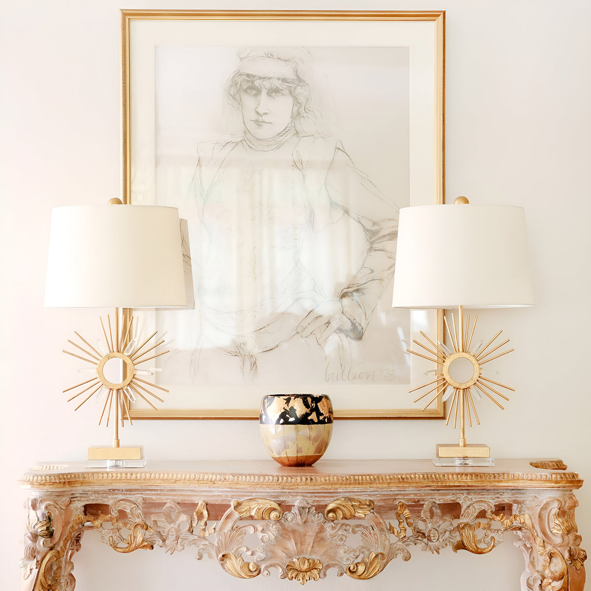 A decorative wooden console table with intricate detailing holds two Sun King 1 Light Table Lamps, gold with ivory shades, alongside a patterned vase. Above the table hangs a framed sketch of a person, all set against a plain white wall.