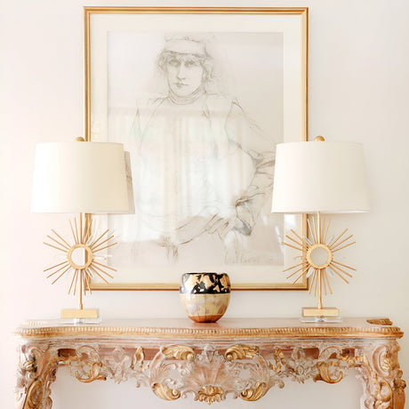 A decorative wooden console table with intricate detailing holds two Sun King 1 Light Table Lamps, gold with ivory shades, alongside a patterned vase. Above the table hangs a framed sketch of a person, all set against a plain white wall.