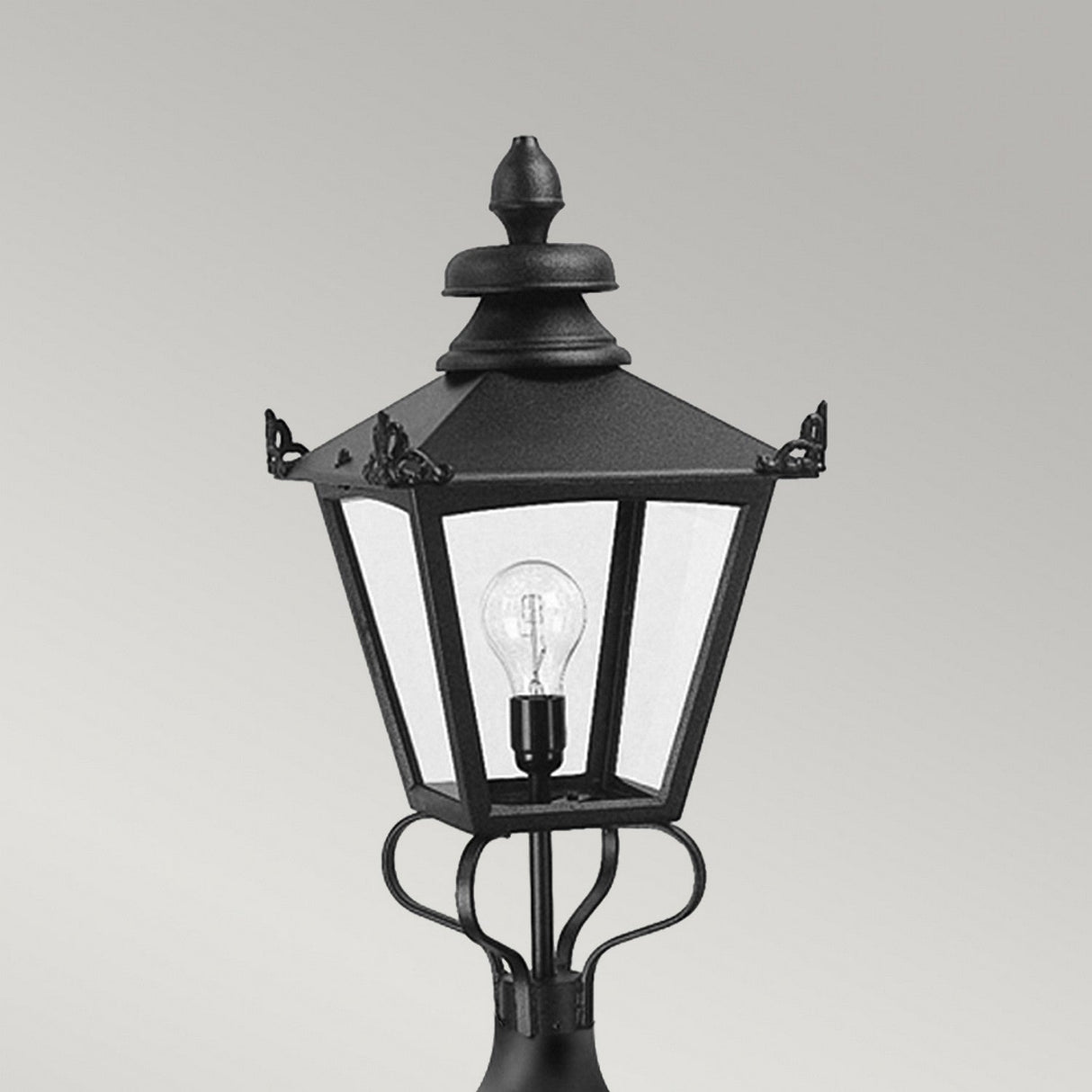 The Grampian 1 Light Pedestal Lantern in black is a vintage-style street lamp crafted from cast aluminum, featuring a clear glass enclosure and a visible light bulb inside, reminiscent of the Victorian lantern style. Set against a neutral gray background, it provides elegant outdoor lighting.
