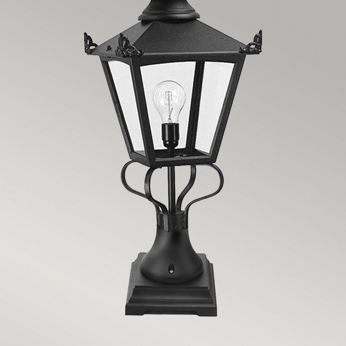 The Grampian 1 Light Pedestal Lantern - Black showcases a Victorian lantern style, made from cast aluminum. It presents a classic design with a clear glass enclosure and an exposed light bulb, mounted on a square base adorned with decorative metalwork—ideal for enhancing your outdoor lighting aesthetic.