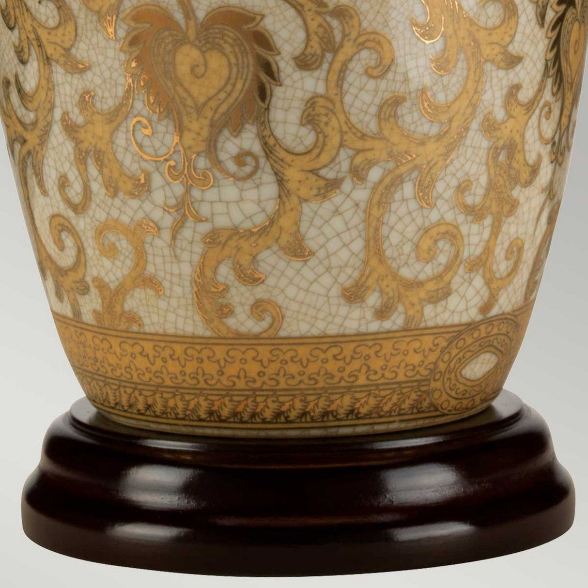 A close-up of the Gold Thistle 1 Light Table Lamp - Gold, with its ornate porcelain base featuring intricate gold floral patterns on a cream background, resting elegantly on a dark wooden stand. The design showcases swirling motifs and heart shapes, all polished to a glossy finish.