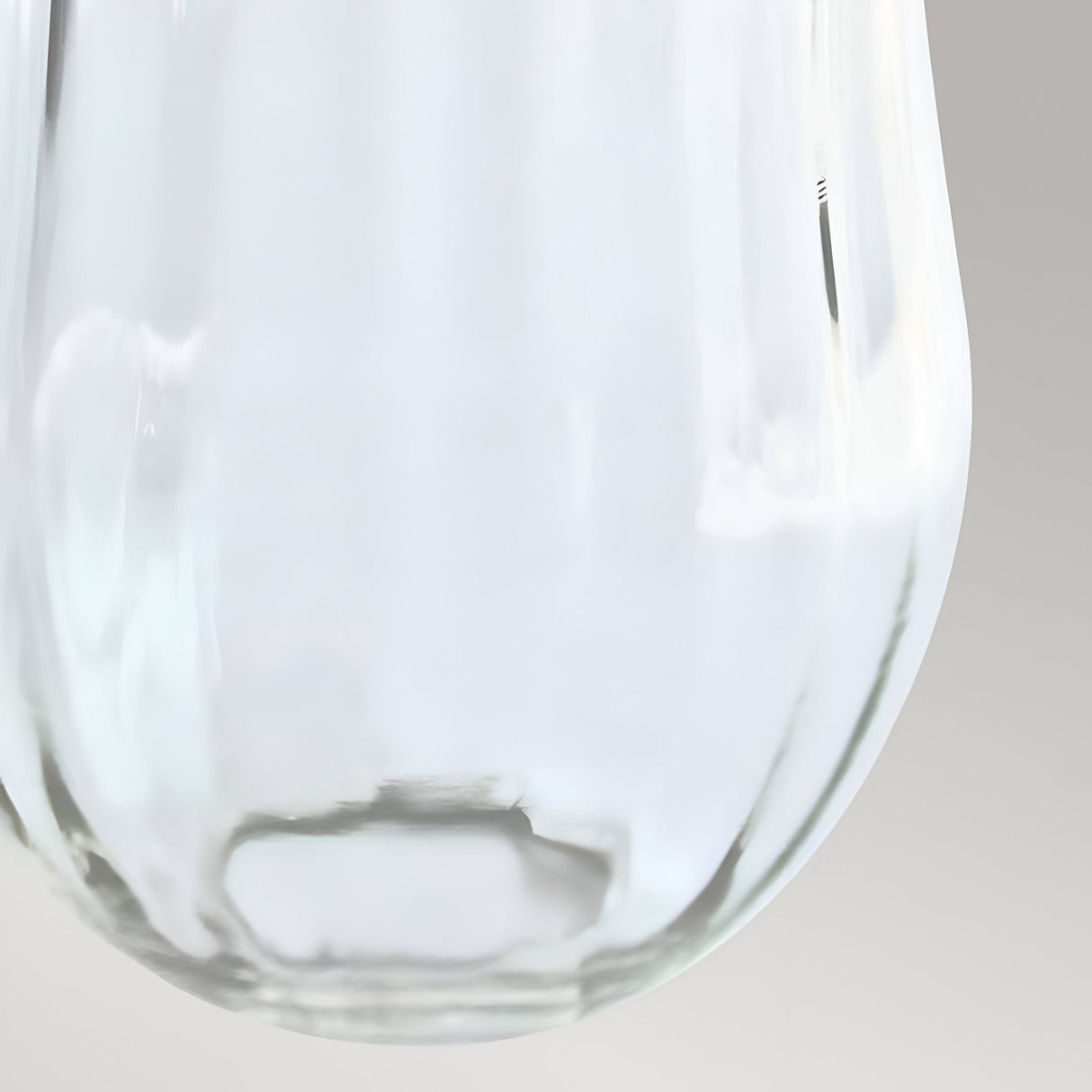 Close-up image of a Glass Shade Optional for Hinkley Cambridge in clear glass against a light gray background. The smooth, reflective surface captures soft light, emphasizing its glossy texture, reminiscent of sophisticated lighting design. No visible light source is lit inside the tall fluted clear glass shade.