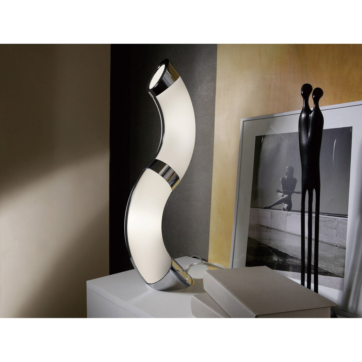 The Guss LED Table Lamp 30K 2700K in polished chrome with white acrylic sits on a white cabinet beside a black abstract sculpture and a framed black-and-white photo, with beige and gray walls in the background.