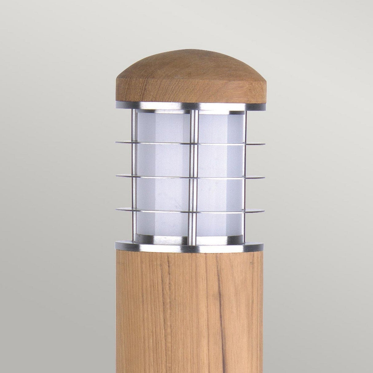 The Poole 1 Light Bollard in light brown showcases a contemporary outdoor design with a cylindrical Malaysian teak base and a Marine Grade 316 stainless steel metal cage surrounding a white frosted glass shade. It is finished with a wooden dome and is set against a simple, light gray background.