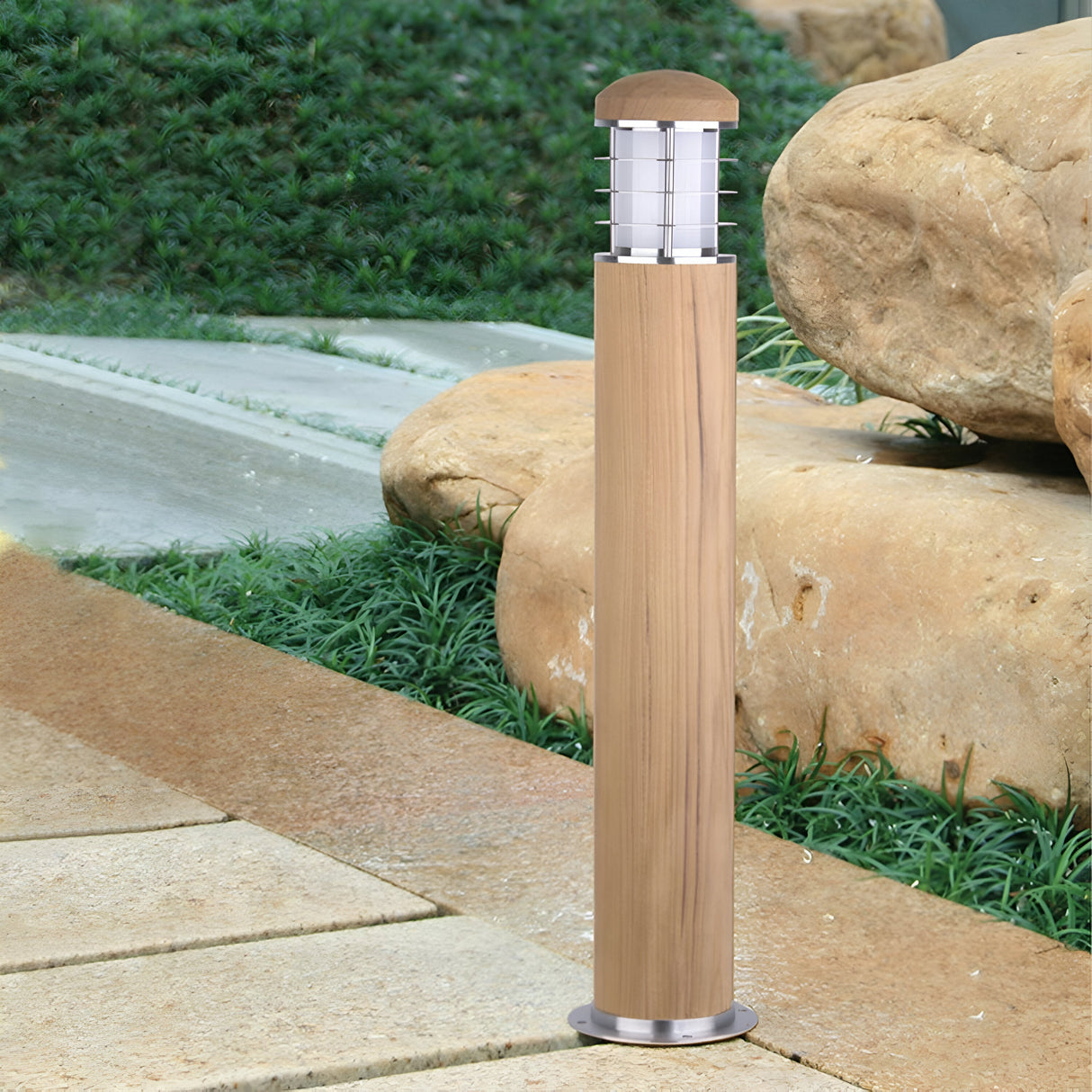 The Poole 1 Light Bollard in light brown, made from Malaysian teak, stands beside a stone path amidst lush greenery and large rocks. Its cylindrical design, enhanced with a Marine Grade 316 stainless steel top and grid-like structure, adds an elegant touch to outdoor lighting.