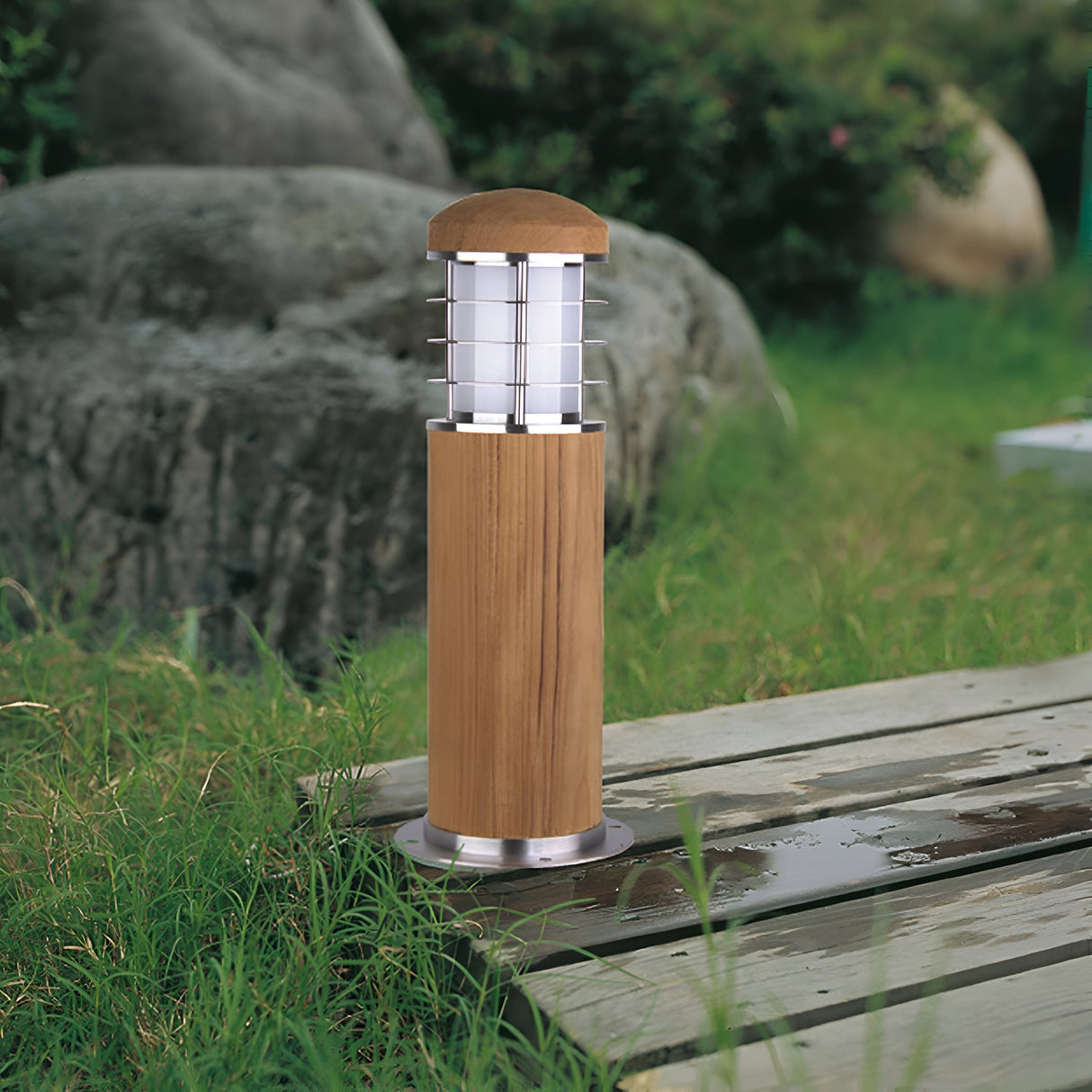 The Poole 1 Light Mini Bollard in light brown, featuring a Marine Grade 316 stainless steel top and gridded cover, is situated on a wooden deck with green grass and rocks in the background.