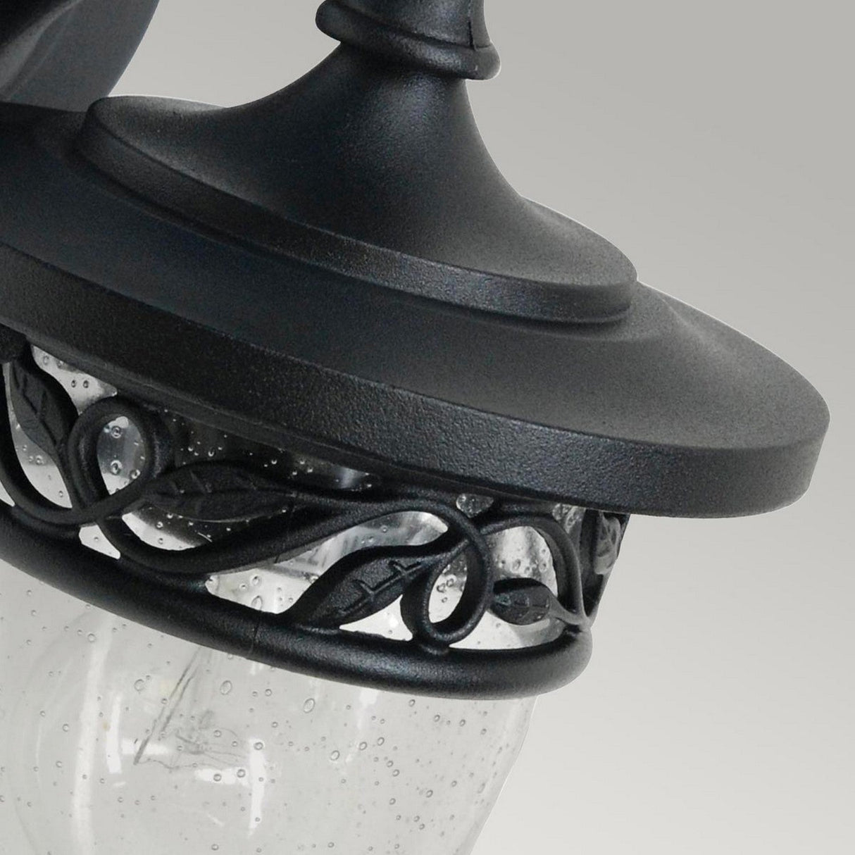 Close-up of the Burford 1 Light Outdoor Wall Lantern in black cast aluminium, showcasing a decorative vine pattern on the glass. Tiny water droplets embellish its surface, adding to its vintage appeal. The lantern's IP44 rating guarantees durability, all set against a simple gray backdrop.