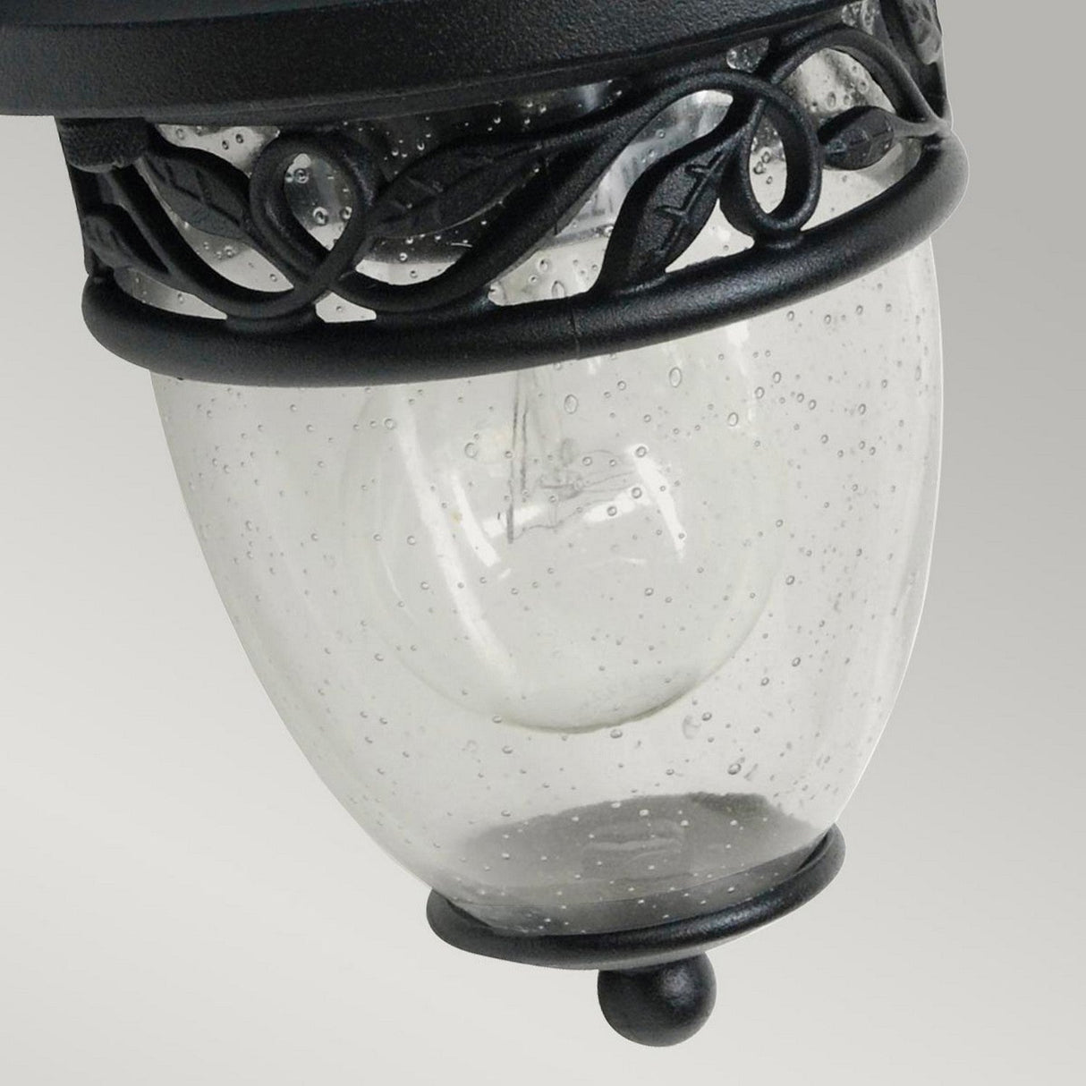 Close-up of the Burford 1 Light Outdoor Wall Lantern in black. The cast aluminum frame showcases leaf-like patterns, and the glass cover is dotted with small air bubbles. This fixture, with its IP44 rating, stands out against a neutral gray background.