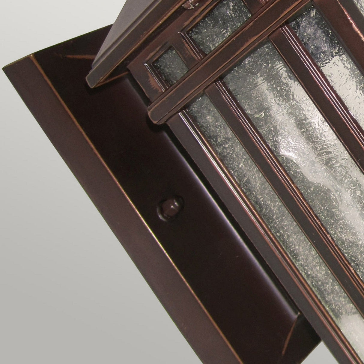 A close-up of the Chedworth 1 Light Outdoor Wall Lantern showcases its durable cast aluminum construction with a bronze finish. The rectangular fixture is enhanced by textured glass panels and is tastefully mounted on a matching bronze wall plate, set against a light gray background.
