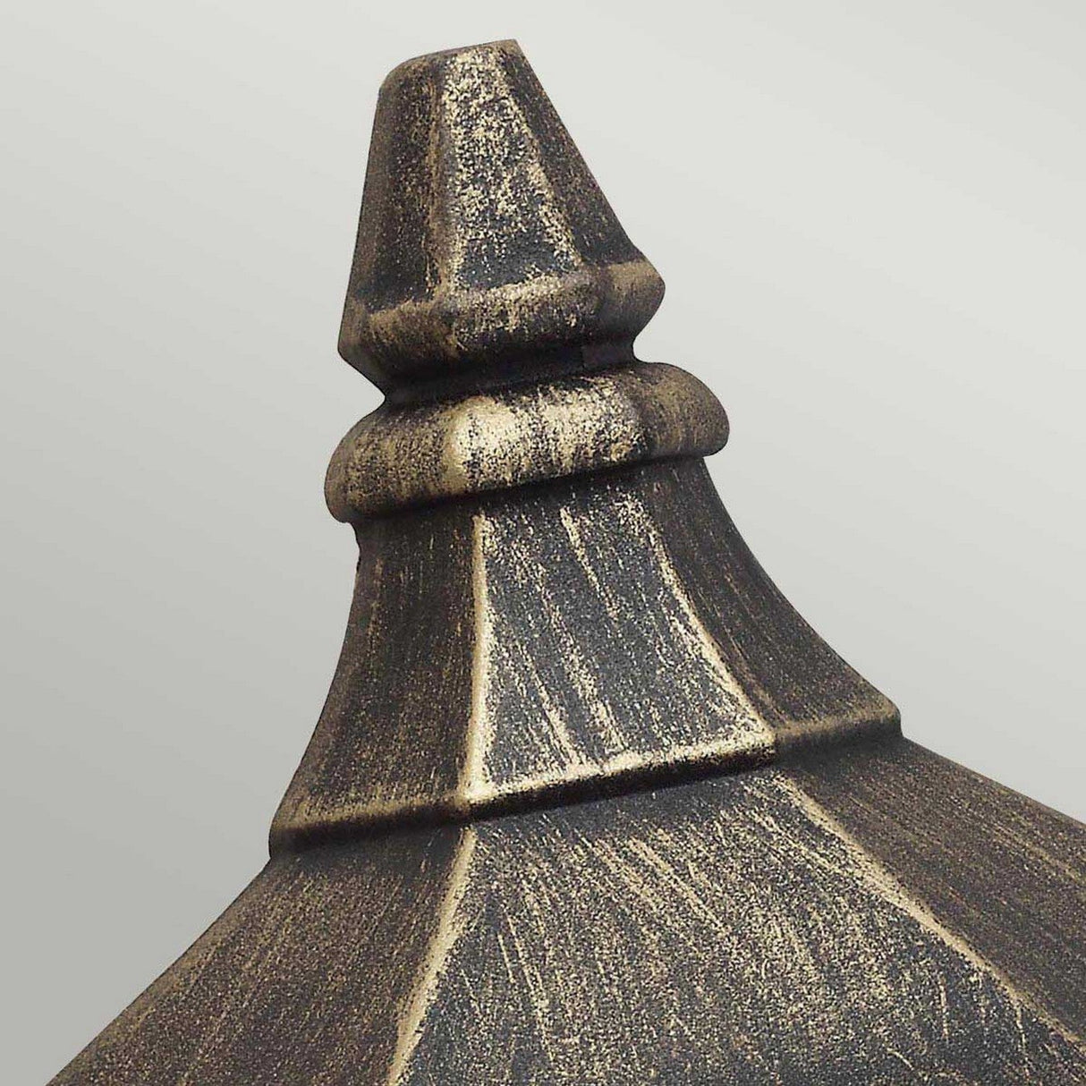 A close-up view of the Cricklade 1 Light Outdoor Half Lantern reveals a weathered, metallic finial with a tapered, multi-tiered design that exudes charm. Its distressed, vintage finish features dark and gold accents on a light gray backdrop, perfectly aligning with its IP44 rated lighting aesthetics.