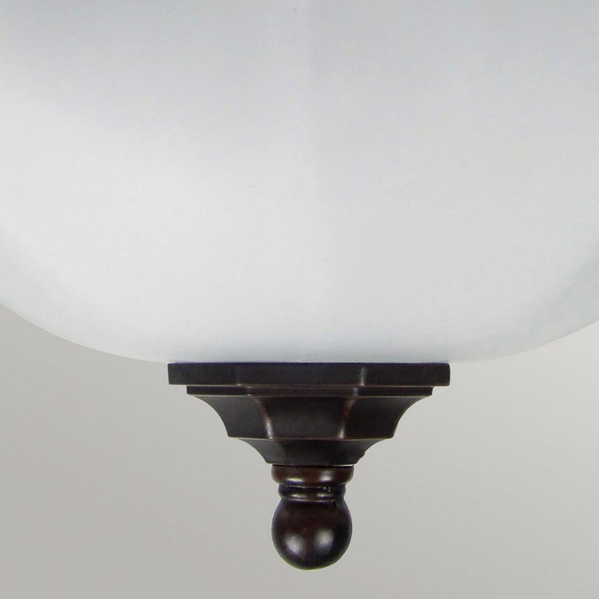 Close-up of an inverted bronze finial with a round base, attached to the bottom of a frosted glass shade, which is part of the IP44 rated Fairford 1 Light Outdoor Flush Lantern in Bronze. The background features a plain, light gray surface.