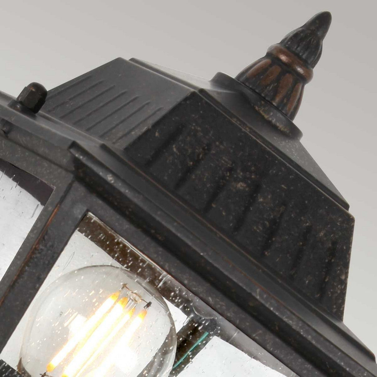 Close-up of the Highnam 1 Light Outdoor Wall Lantern in a vintage-style metal design with a weathered bronze finish. The cast aluminum structure features a peaked top and decorative finial, while glass panels reveal an illuminated bulb inside, adding rustic charm.