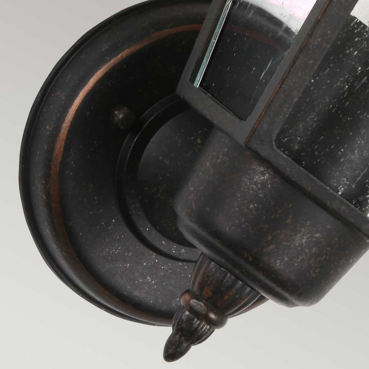 Close-up of the Highnam 1 Light Outdoor Wall Lantern in a weathered bronze finish, showcasing a cylindrical glass enclosure and decorative top and bottom finials. Mounted on a circular base, the cast aluminum metal enhances its rustic charm with a weathered texture.