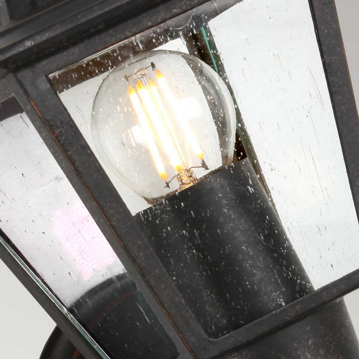 Close-up of a glowing bulb inside the Highnam 1 Light Outdoor Wall Lantern in a weathered bronze finish. The cast aluminum frame beautifully complements the glass panels speckled with rain droplets, casting a cozy, warm glow against the gray sky.