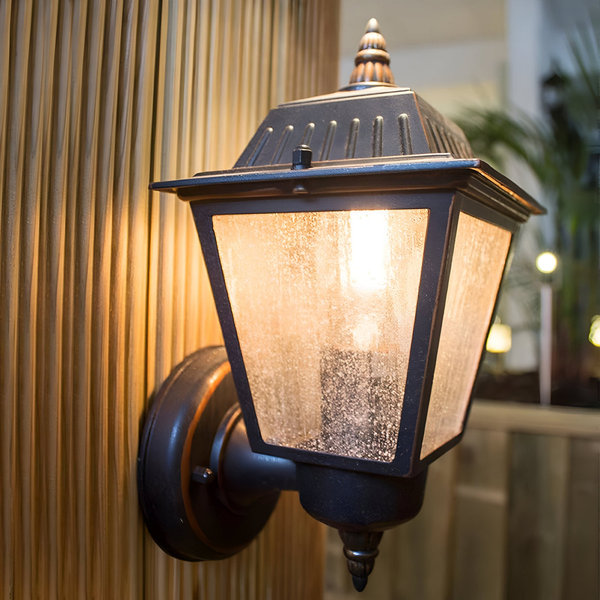 The Highnam 1 Light Outdoor Wall Lantern in weathered bronze, made from cast aluminum, gently lights up the area. Its frosted glass cover complements a wooden wall, creating a warm and timeless atmosphere.