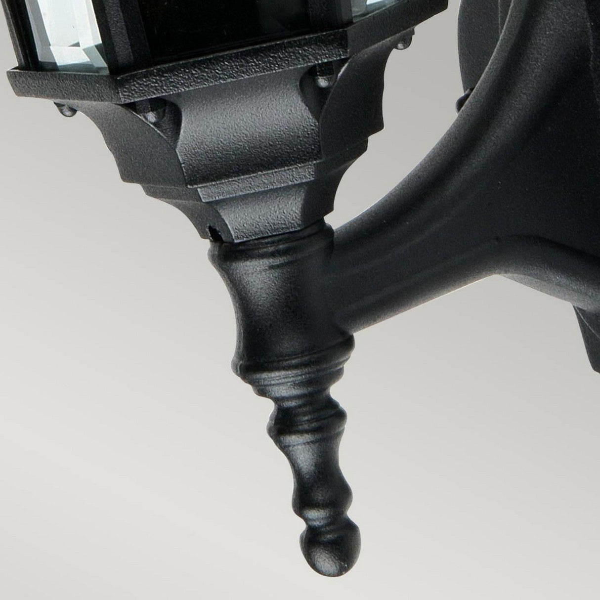 Close-up of the Ledbury 1 Light Wall Lantern - Black bracket, crafted from cast aluminum with a decorative spindle. The background is a smooth, light gray surface. This outdoor lantern boasts a classic, ornate style, perfect for enhancing your decorative exterior lighting setup.