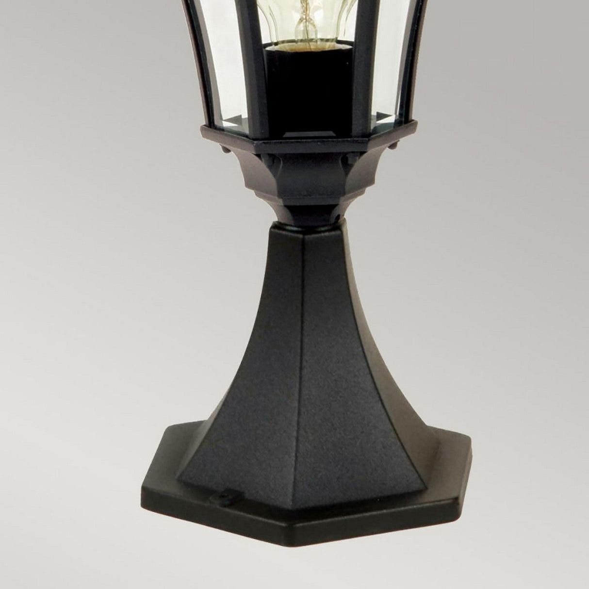 A close-up of the Ledbury 1 Light Pedestal Lantern in black, crafted from cast aluminium with a hexagonal base and clear glass panels revealing a light bulb inside. The plain light gray background accentuates this decorative piece of exterior lighting.