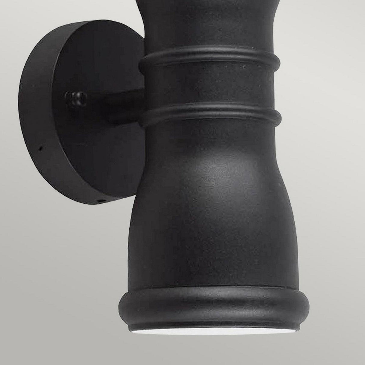 Close-up of the Malvern 2 Light Up & Down Lantern in black, a modern-style wall-mounted outdoor light fixture made from cast aluminum. This cylindrical piece features two horizontal ridges near the top and a matte finish. It is attached to a round mounting plate, offering decorative exterior lighting.