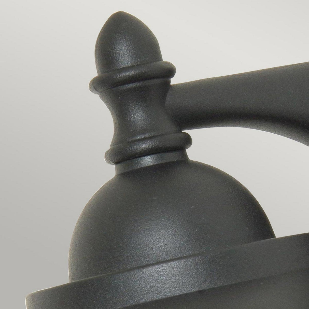 A close-up of the Sandown 1 Light Outdoor Wall Lantern - Black, crafted from cast aluminium, showcases its rounded top finial with a pointed tip. The smooth, matte finish beautifully contrasts against the plain, light-colored background.