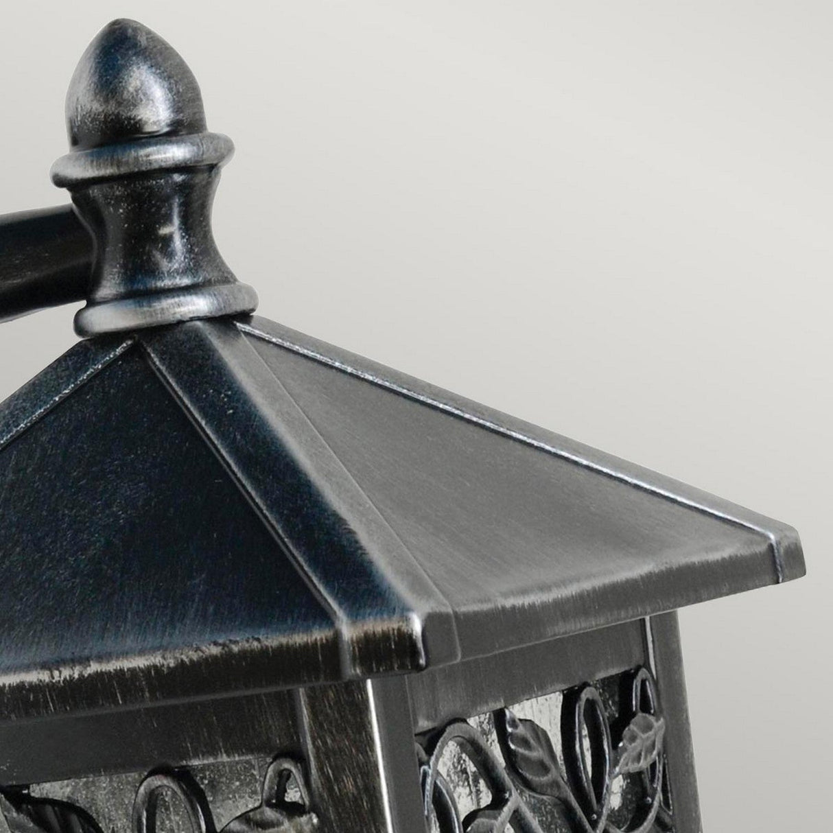 Close-up of the Winchcombe 1 Light Outdoor Wall Lantern - Black, crafted from cast aluminum with a pointed top and intricate leaf patterns adorning its metal frame. This IP44 rated fixture features a dark, metallic finish, set against a plain, light gray background.