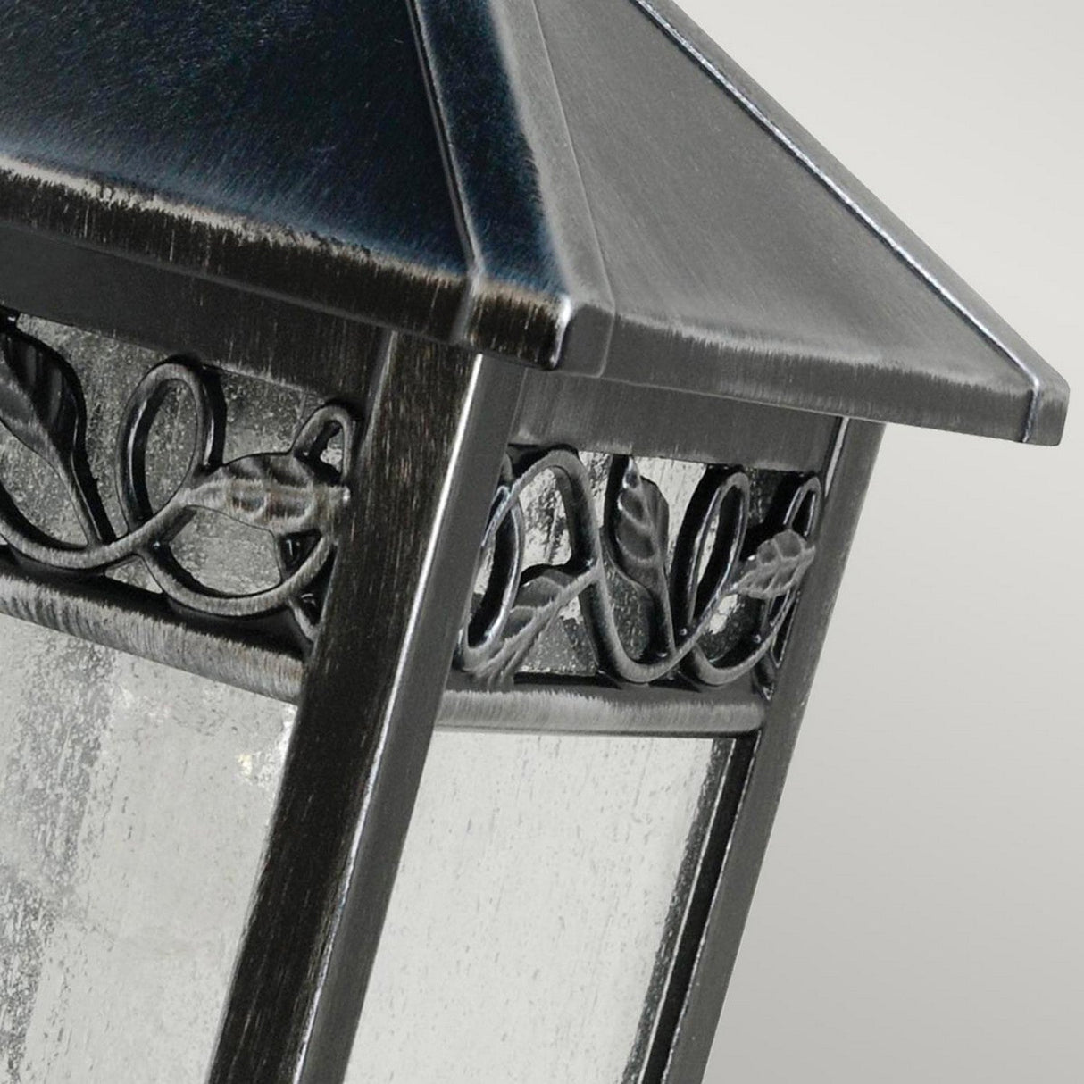 The Winchcombe 1 Light Outdoor Wall Lantern - Black features a close-up view of its durable aluminum construction and decorative vine pattern along the sides. This elegant lantern boasts a textured glass surface and a pointed roof, ideal for IP44-rated lighting setups against a neutral gray background.