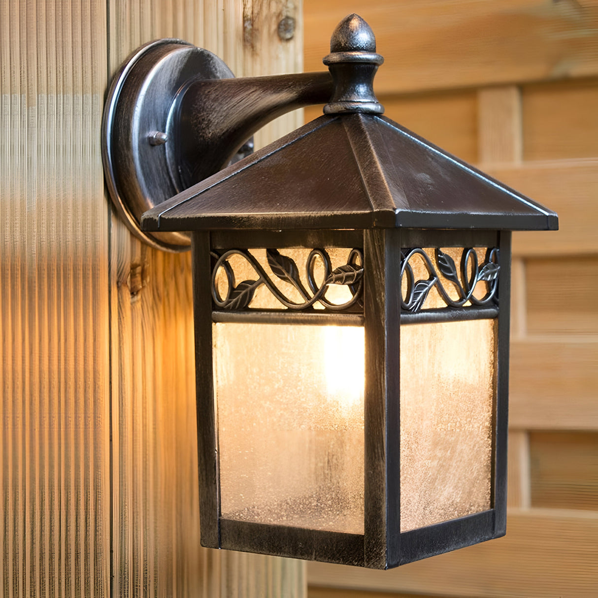 The Winchcombe 1 Light Outdoor Wall Lantern - Black, crafted from cast aluminum with a metal frame and frosted glass panes, is mounted on a wooden wall. This IP44 rated fixture casts a warm glow and features decorative leaf patterns on its upper section.