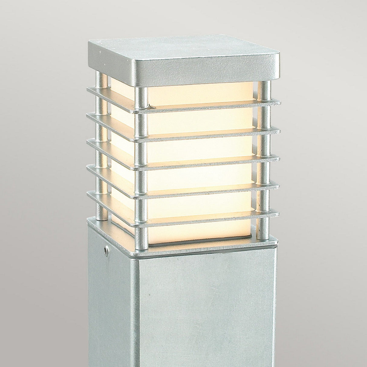 The Halmstad Large Bollard - Silver is a modern outdoor lamp post made from galvanized steel, showcasing a rectangular shape with a grid-like metal frame. This fixture casts a soft, warm glow against a plain gray background.