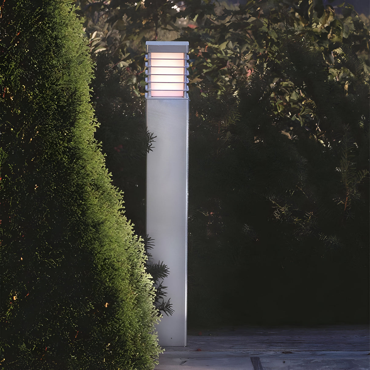 A contemporary Halmstad Large Bollard - Silver, featuring a glowing light at the top, stands beside lush green bushes. Made from galvanized steel, it enhances the garden's ambiance, perfectly complementing the dense foliage with its sophisticated outdoor lighting design.