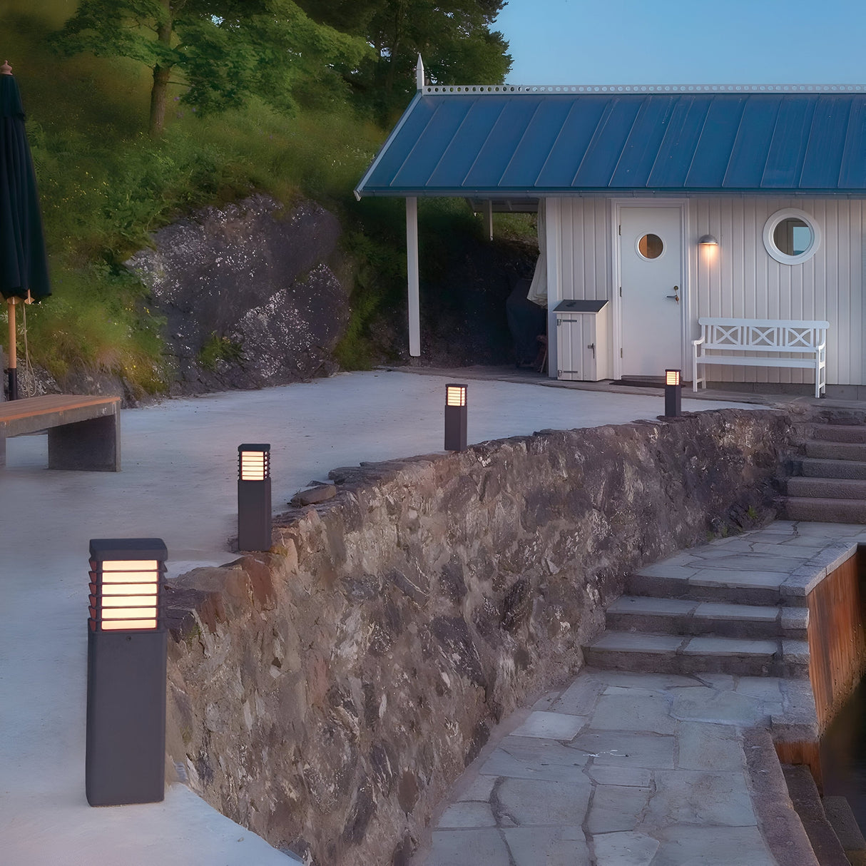 A modern landscaped outdoor space highlights a small, white, wooden house with a blue roof. The area features the Halmstad 1 Light Medium Bollard in black, elegantly lining a stone wall beside steps leading to the house, set against a backdrop of trees and rocks under a clear evening sky.
