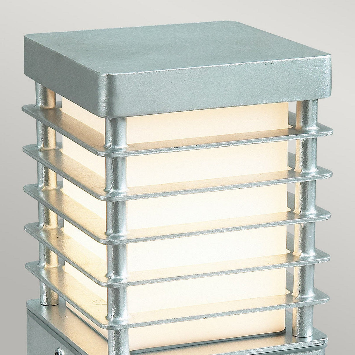 The Halmstad Medium Bollard - Silver is a contemporary outdoor pillar light featuring a square design. Made from galvanized steel, it has horizontal bars encircling a frosted glass panel that emits a soft, warm glow. This stylish outdoor lighting fixture stands gracefully against a neutral gray background.