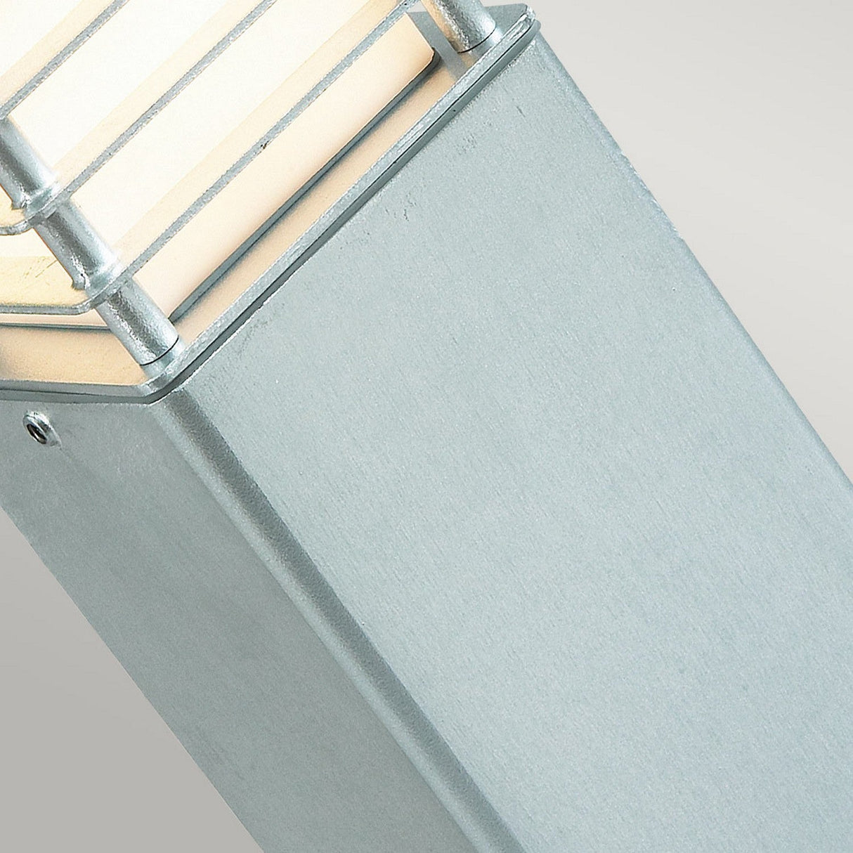 Up-close view of the Halmstad Medium Bollard - Silver, a contemporary outdoor light designed with a square shape and horizontal metallic bars across the top. Made from galvanized steel, it boasts a brushed finish and emits a warm glow, making it perfect for elegant outdoor lighting.