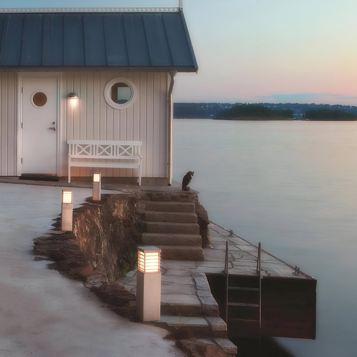 A small white boathouse with a circular window and a bench sits by a calm lake at sunset. A cat relaxes on the stone steps leading to the water, where Halmstad Medium Bollard - Silver lamps gently illuminate the path. A distant shoreline is visible across the lake.