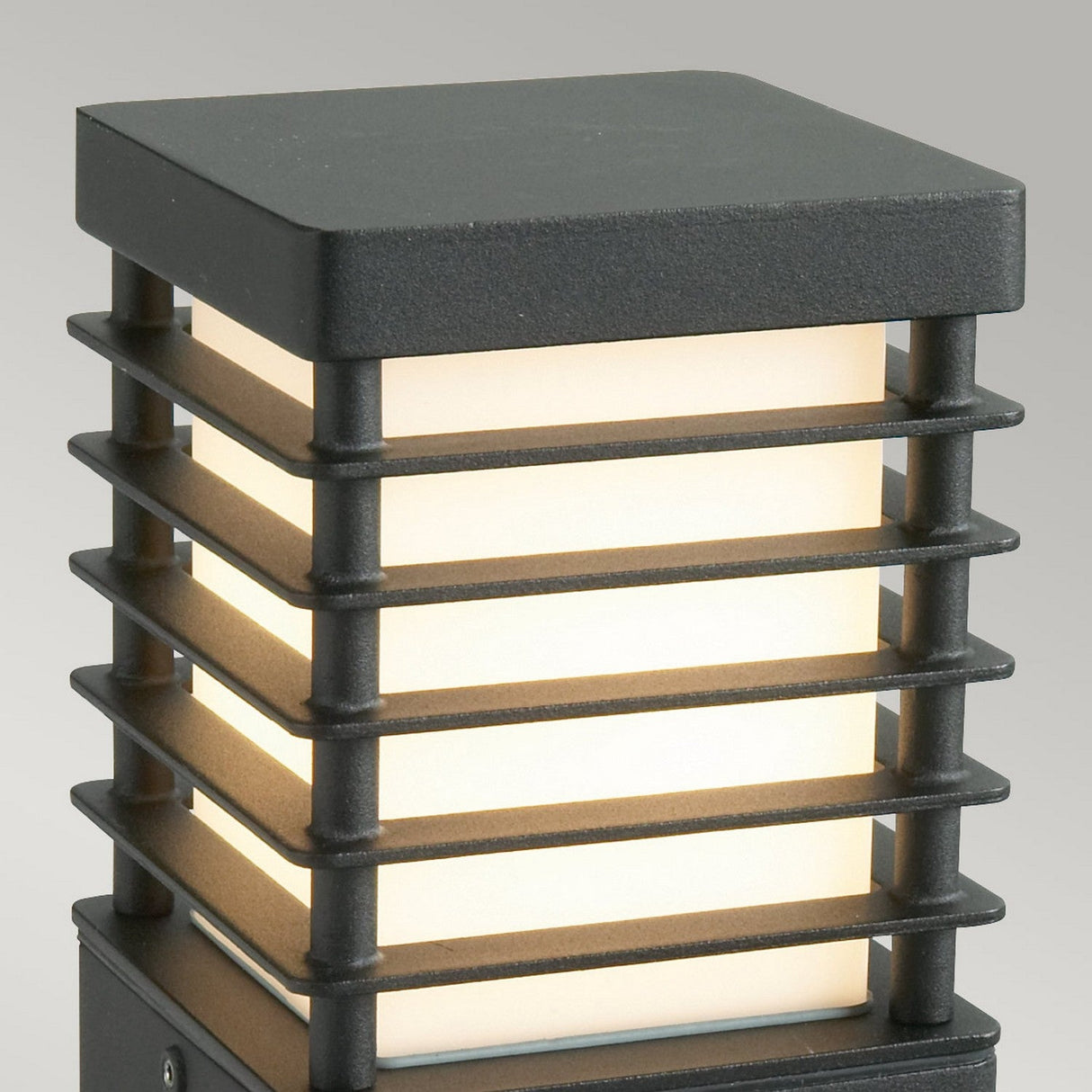 The Halmstad Small Bollard Black showcases a modern rectangular design with horizontal metal bars encasing a frosted glass center, all in a sleek black finish. It emits a warm glow against a neutral gray backdrop, making it perfect for enhancing any contemporary setting.