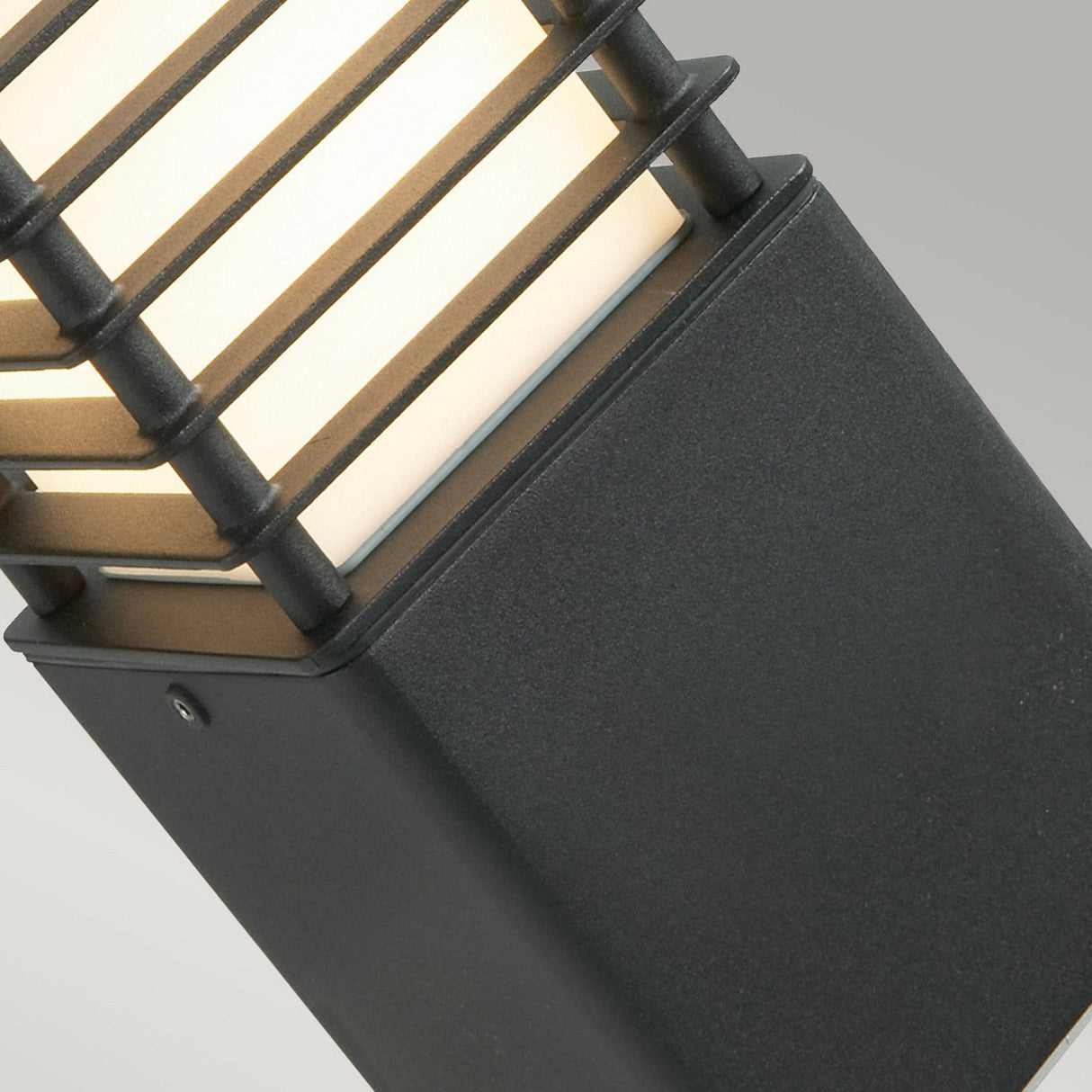 Close-up of the Halmstad Small Bollard Black, showcasing sleek outdoor lighting. This modern fixture features a rectangular design with horizontal metal slats above a solid black base, creating a contemporary look. The black finish beautifully complements the soft glow against the light gray background.