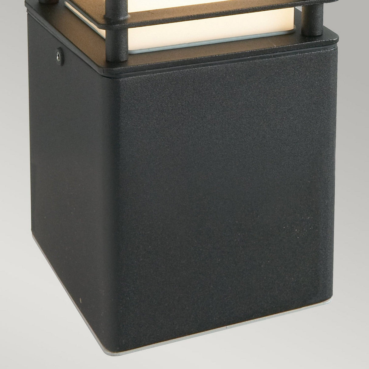 A close-up of the Halmstad Small Bollard in black, featuring a frosted glass panel on top to emit a gentle glow. Its modern, minimalist design makes it perfect for mounting on a plain grey wall to enhance outdoor lighting aesthetics.