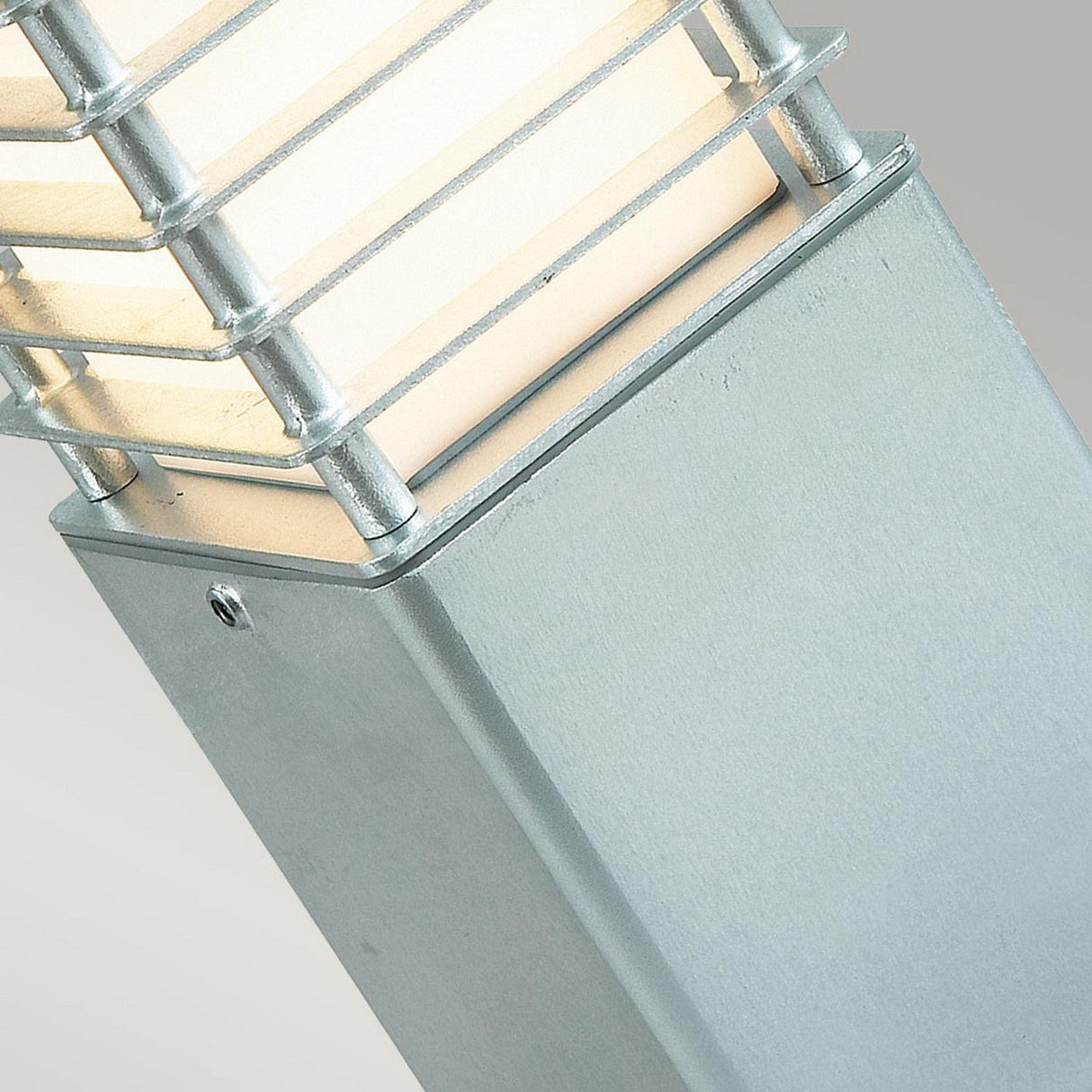 The close-up image highlights the Halmstad Small Bollard in Silver, featuring a modern design with a rectangular galvanized steel base and a frosted glass top. Horizontal metal bars wrap around the glass to elegantly diffuse light. Rated IP65, it is built to endure various weather conditions, against a plain light gray background.