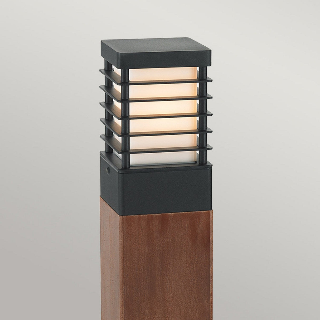 The Halmstad 1 Light Medium Wooden Bollard in black is a contemporary outdoor lamp post ideal for pathway lighting. It boasts a wooden bollard base and a square metal lamp with a grid design that emits a soft, warm glow.
