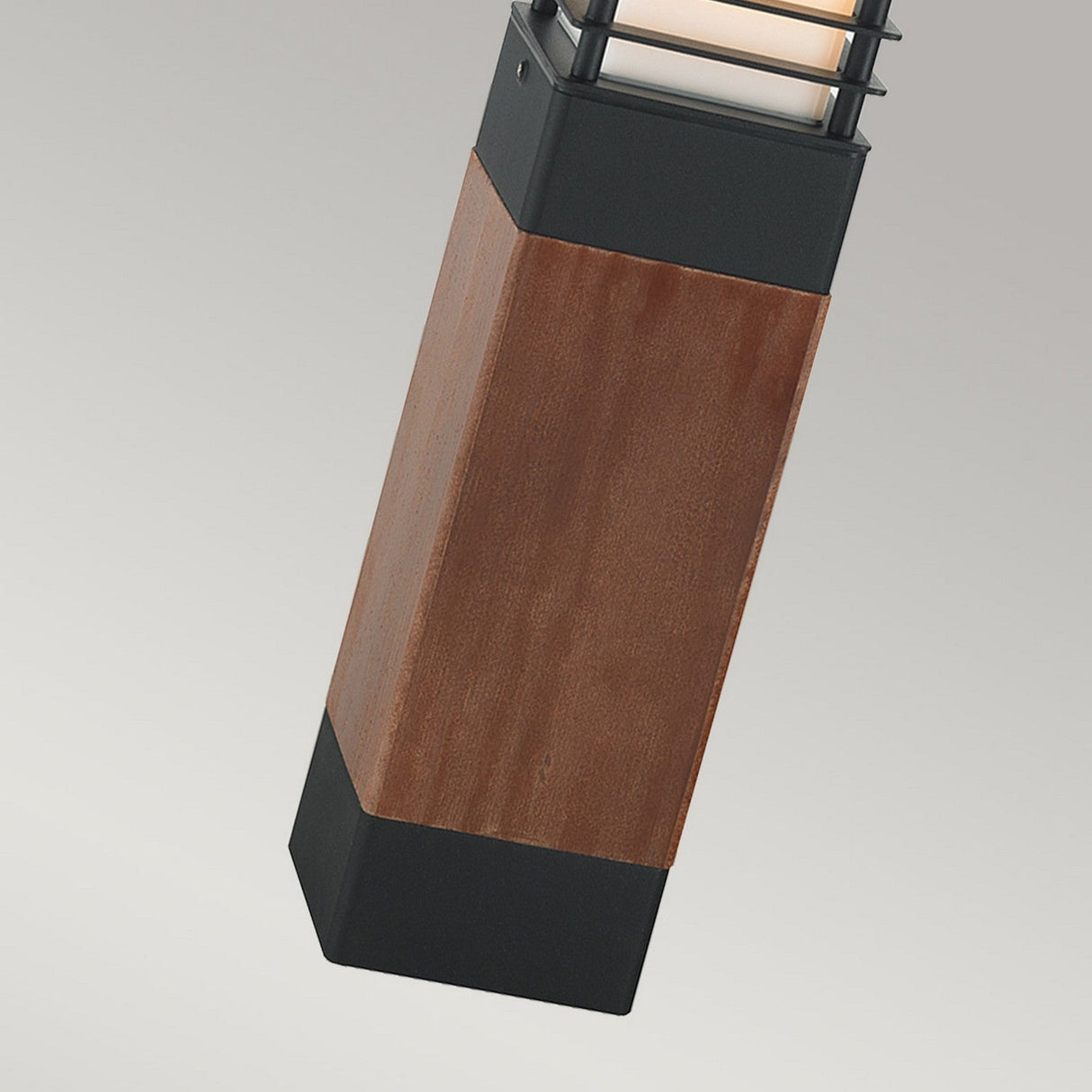 A close-up of the Halmstad 1 Light Medium Wooden Bollard - Black highlights its modern cubic design, reminiscent of charming outdoor lighting. The fixture showcases a wooden central section, bordered by sleek black metal edges and is topped with a metal grid surrounding the light source. The background is a simple gray.