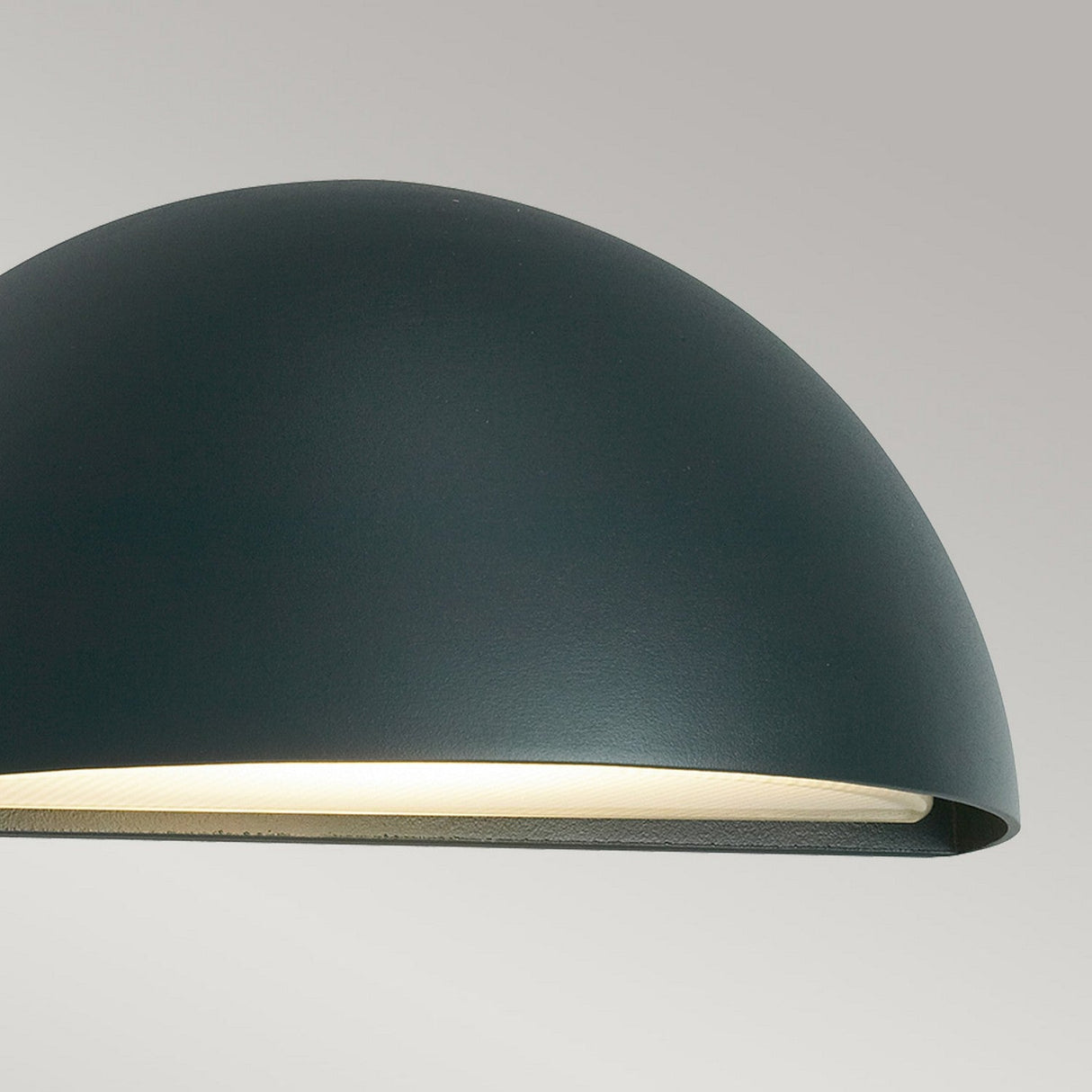 A close-up of the Halden Outdoor 1 Light Down Wall Light in graphite showcases its modern dome-shaped design that casts a soft, warm glow against a light gray backdrop. Its minimalist style accentuates the smooth, curved surface of the lampshade, adding an elegant aesthetic.