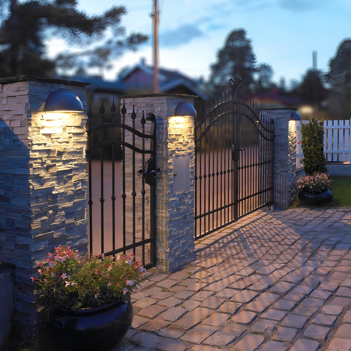 A black wrought iron gate with stone pillars is adorned with a Halden Outdoor 1 Light Down Wall Light in graphite, its IP65 rating ensuring a soft glow over the cobblestone driveway. Potted plants brimming with pink flowers flank the entrance, while a wooden fence and trees provide a picturesque backdrop under the twilight sky.