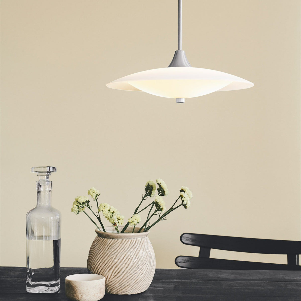 Baroni Aluminium Pendant Light 40cm in Opal, a wide shallow white disc with a glowing opal glass bowl beneath, shown lit above a dining table with a vase and decanter.