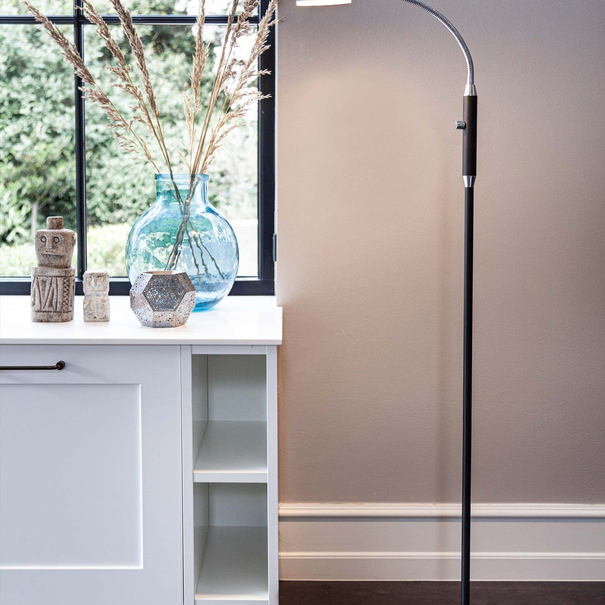 Vegas LED Floor Lamp 7W in black, shown beside a window with a slim black column, chrome gooseneck arm and flat head, placed near a white cabinet with decorative vases.
