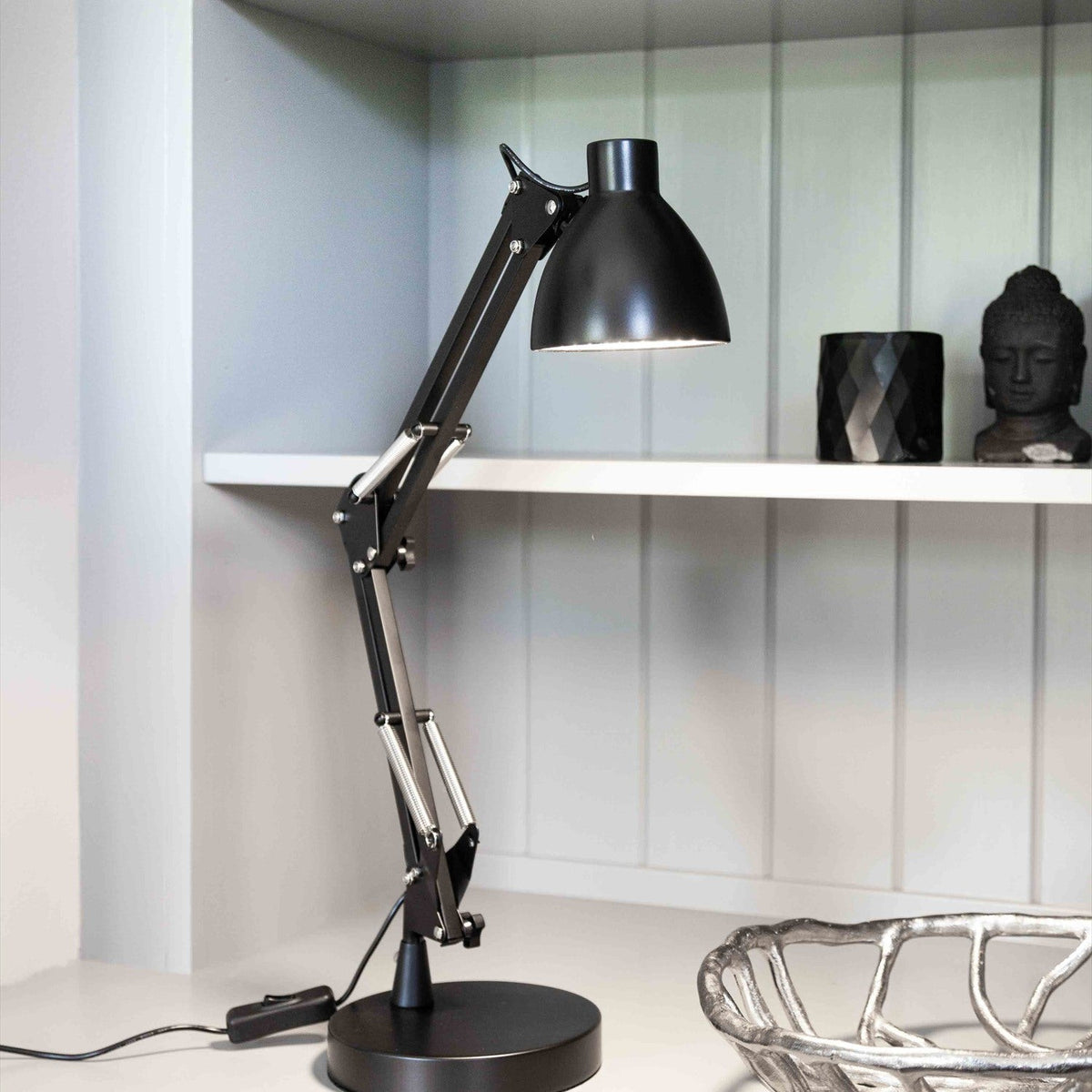 Bronx LED Table Lamp in black, an adjustable articulated desk lamp with folding arms and cone-shaped head, shown on a shelf beside dark decor ornaments.
