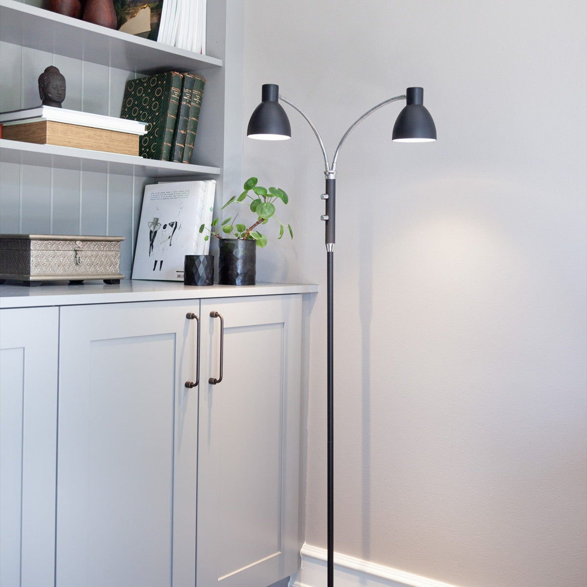 Hudson 2 Light Floor Lamp in black with slim column, chrome gooseneck arms and adjustable black cone heads, shown beside a grey shelving unit and cabinets.
